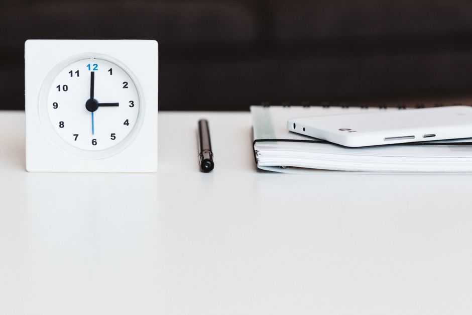 A minimalist office desk setup with a clock, pen, smartphone, and notebook, conveying time management and organization.